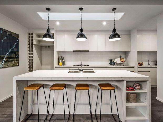 Contemporary white kitchen with a large waterfall-edge island, four timber-topped black metal bar stools, integrated white cabinetry, open shelving, dark tiled flooring, and three black pendant lights suspended beneath a large rectangular skylight. A narrow wine-bottle storage rack and built-in shelving flank the cabinetry.