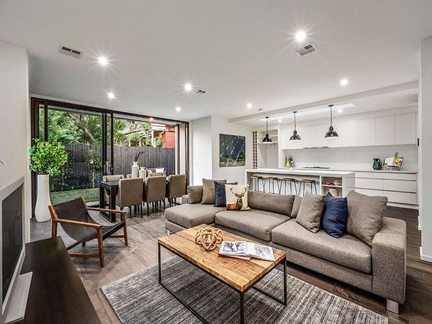 Open-plan contemporary living, dining and kitchen interior with a large sectional sofa, timber coffee table, dining setting, kitchen island and extensive glazed doors opening to a planted outdoor area. The room has white walls and ceiling, dark timber flooring, neutral furnishings, pendant lighting and integrated cabinetry.