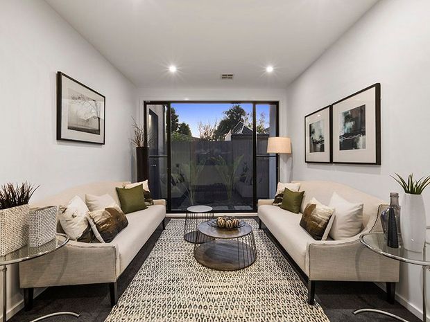 A compact, bright contemporary living room with two facing cream sofas, patterned cushions, round nested coffee tables, a large geometric rug, framed monochrome artwork, and potted plants. Full-height glazed doors open toward a small enclosed outdoor area, while recessed ceiling lights and a floor lamp provide layered illumination.