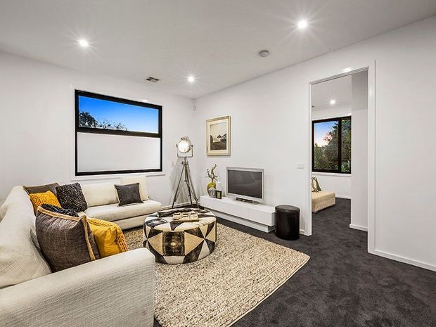 Bright contemporary living room with a large cream sectional sofa, patterned cushions, a geometric round coffee table, textured area rug, wall-mounted television and tripod floor lamp. White walls and recessed ceiling lights contrast with dark charcoal carpet. An open doorway reveals part of an adjacent bedroom or sitting area.