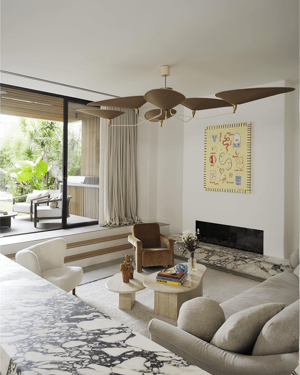 Bright, refined living room with a large sculptural multi-arm ceiling fan, neutral upholstered seating, a stone coffee table, and a wide linear fireplace set into a pale wall with a rough stone hearth. Floor-to-ceiling sliding glass doors open to a planted outdoor terrace, with layered steps and full-height curtains. A large veined stone surface occupies the foreground, likely an adjacent kitchen or counter edge. The room combines organic textures, warm neutrals, and mid-century-inspired furnishings.