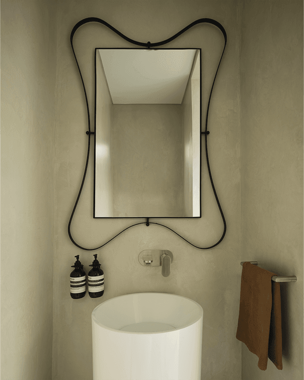 A compact, moody bathroom vanity area with a freestanding white vessel basin, wall-mounted brushed metal tap, rectangular mirror, and sculptural black cord or metal frame surrounding the mirror. The walls have a textured warm taupe plaster finish, with dark soap dispensers and a brown hand towel adding contrast.
