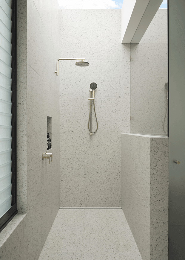 A minimalist walk-in shower enclosed by pale speckled stone-look slab surfaces, with matching floor and half-height partition. Brushed brass shower fittings contrast with a dark hand-shower control. A narrow window with a dark frame sits to the left, while a skylight or open ceiling detail brings in daylight.