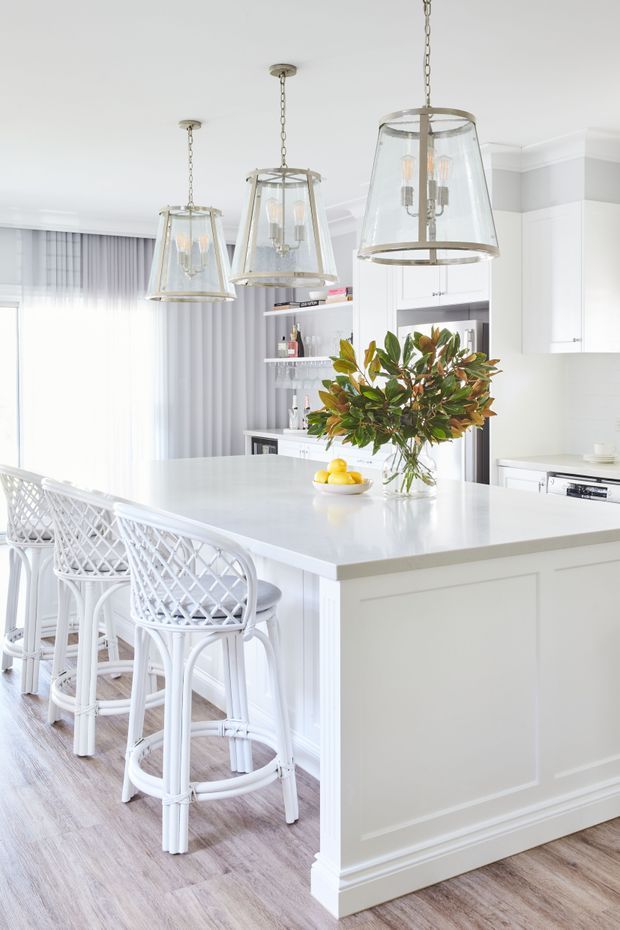 Bright, airy white kitchen featuring a large paneled island with a pale stone benchtop, three glass-and-metal pendant lights, white cabinetry, open shelving, woven white bar stools, and light timber-look flooring. A leafy arrangement and bowl of lemons provide natural colour accents. Soft daylight enters through sheer curtains.