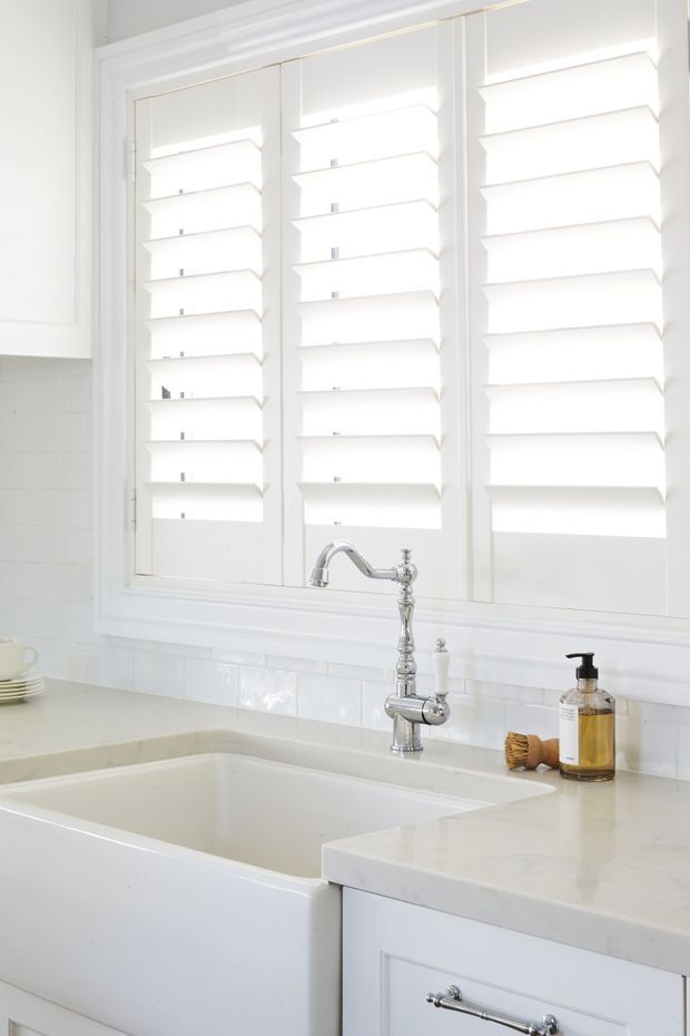 Bright, white traditional-style kitchen sink area featuring a deep apron-front farmhouse sink, pale countertop, white tiled backsplash, white fixed cabinetry, and a large three-panel window fitted with white plantation shutters. A polished chrome bridge-style tap and small countertop accessories sit beside the sink.