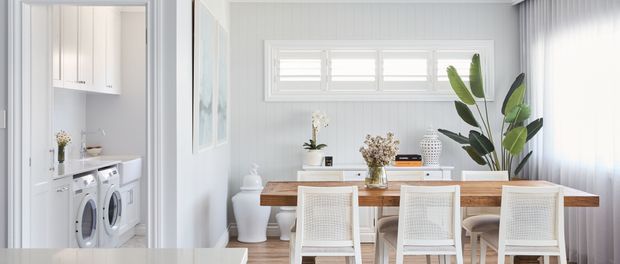 Bright white dining room with a timber dining table, three woven-back dining chairs, a large leafy indoor plant, and white vertical wall panelling. A compact laundry area with white cabinetry and front-loading appliances is visible through the opening on the left.