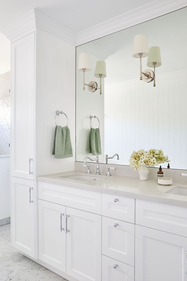 Bright, refined bathroom vanity with extensive white paneled cabinetry, a pale stone benchtop, chrome hardware and tapware, framed wall mirror, twin wall sconces, and a marble-look floor. Soft green towels and a small floral arrangement provide subtle colour accents.