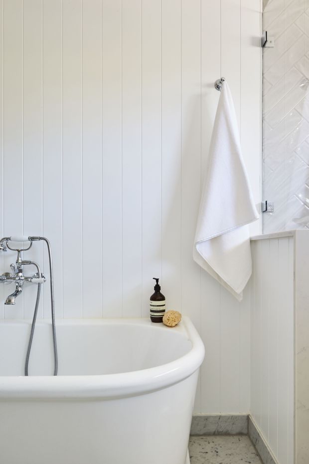 Bright, compact bathroom featuring a freestanding white bathtub, chrome wall-mounted bath tap and shower fittings, white vertical wall panelling, a white textured tiled shower enclosure, and a small white towel on a chrome hook. The floor is finished in grey-white stone-look tile, with minimal dark soap and natural sponge accessories on the tub edge.