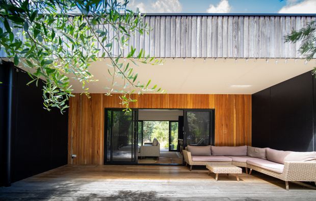 A contemporary outdoor living terrace framed by black walls, with a warm timber-clad rear facade, large sliding glass doors, and a low-profile upholstered sectional lounge. A projecting upper volume has a pale smooth soffit and vertically ribbed metal cladding, while mature leafy branches provide shade across the space.