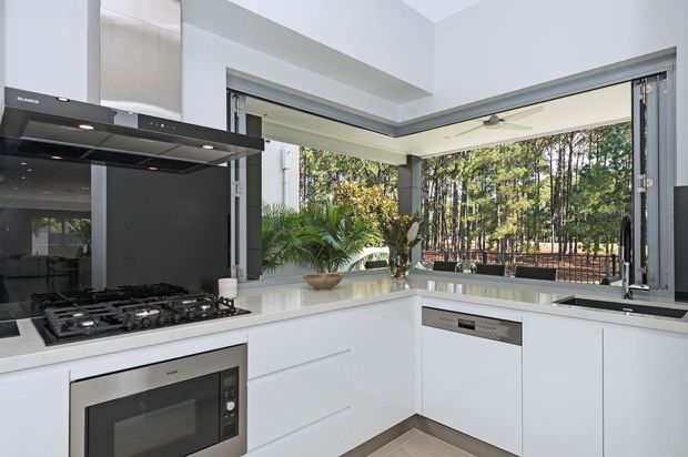 Bright contemporary kitchen with white handleless cabinetry, a pale stone benchtop, integrated appliances, black glass splashback and a wide corner opening with folding or bi-fold windows overlooking trees. The room has a clean, open feel with dark aluminium window frames and timber-look flooring.