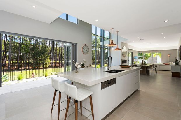 Bright open-plan contemporary kitchen and living area with a large white island, integrated cooktop and sink, white cabinetry, timber-legged bar stools, pendant lights, and expansive floor-to-ceiling glazing overlooking a wooded outdoor setting. A dining and living area extends beyond the kitchen.