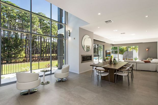 Bright, spacious open-plan living and dining area with a dramatic double-height glazed wall overlooking a wooded landscape. The room features large-format grey tile flooring, a neutral white and grey palette, contemporary built-in fireplace, sculptural lounge chairs, a substantial timber dining table, and extensive indoor-outdoor glazing.