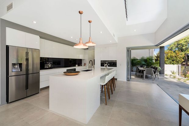 Bright, open-plan contemporary kitchen with extensive white flat-panel cabinetry, a large white island with bar seating, stainless-steel appliances, a glossy black splashback, pale tiled flooring, and copper pendant lights. Large glazed sliding doors open directly to a furnished outdoor patio, while a ceiling void or skylight brings additional natural light into the space.