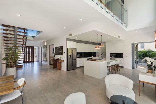 Bright, open-plan contemporary interior featuring a white kitchen with central island and breakfast-bar seating, adjoining living and dining areas, a floating timber-and-metal staircase, large-format grey floor tiles, and a double-height void with glass balustrade. The space has abundant daylight, recessed lighting, white walls, and timber accents.