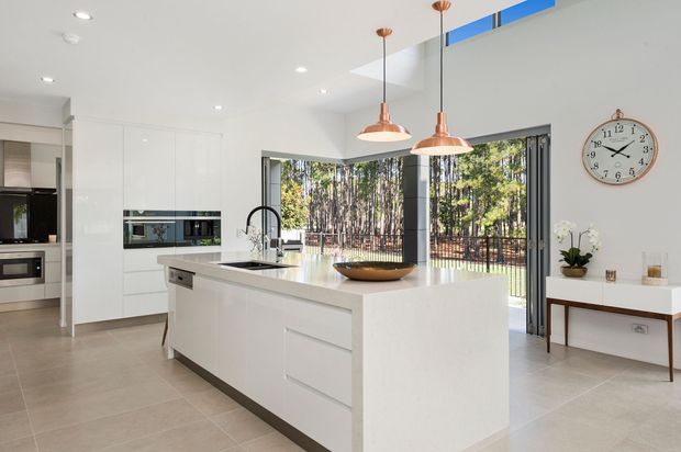 Bright contemporary open-plan kitchen with a large white island, integrated white cabinetry, pale tiled flooring, copper pendant lights, and full-height glazing opening to a deck and wooded landscape. A small console and wall clock sit along the adjacent wall.