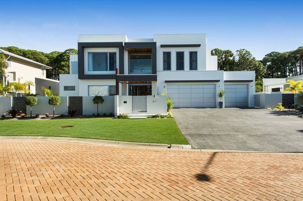 Street-facing view of a contemporary two-storey suburban residence with a crisp white rendered facade, flat roof forms, projecting rectangular volumes, dark-framed glazing, a recessed timber-toned entry, and an integrated double garage. The home is set behind a manicured lawn and low boundary walls, with a broad paved driveway and brick-paved street in the foreground.