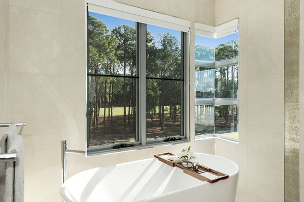 A bright contemporary bathroom featuring a freestanding white bathtub positioned beside large corner windows. The windows provide expansive views of a wooded landscape, while pale stone-look wall surfaces, slim dark window frames, and strong natural sunlight create an airy, serene setting. A timber bath tray with small plants and accessories rests across the tub.