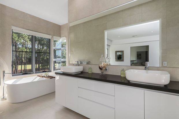 Bright contemporary bathroom with a freestanding white bathtub beside large black-framed windows, a wide double vanity with white vessel basins and dark benchtop, pale large-format wall and floor tiles, and a full-width mirror. Trees and a pool-like outdoor area are visible through the glazing.
