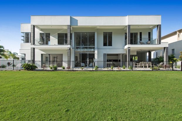 Front exterior view of a large contemporary multi-storey residential building with a crisp white rendered facade, flat rooflines, broad balconies, extensive glazing, black metal balustrades, and ground-level outdoor living areas. The building is viewed across a broad lawn under a clear blue sky.