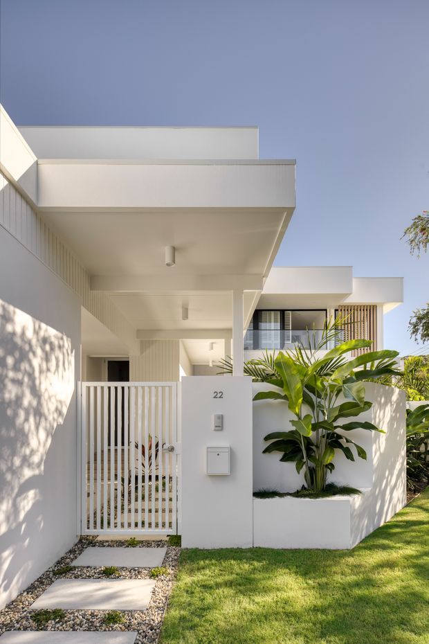 A bright, contemporary residential townhouse entrance viewed from the landscaped front garden. The white rendered facade has strong horizontal roof projections, recessed entry walkways, a slim white metal security gate, minimalist wall-mounted letterbox and intercom, and a raised planter with tropical foliage. A second-storey balcony or window zone with vertical timber screening is visible behind the entry canopy. The composition is clean, geometric, and sunlit against a clear blue sky.