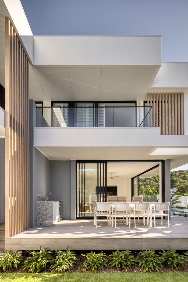 A bright contemporary two-storey residential exterior showing a covered outdoor dining terrace, upper balcony with clear glass balustrade, white rendered projecting roof and balcony volumes, black-framed glazing, and vertical timber screening. Lush planted garden beds sit in front of the raised deck.