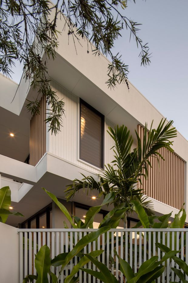 A close-up exterior view of a contemporary multi-level residence, framed by lush tropical planting. The building features clean white rendered volumes, projecting balconies, dark-framed glazing, vertical timber-look screening and slatted fencing, with warm recessed lighting visible beneath the overhangs.