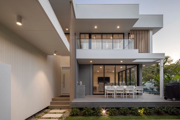 Contemporary two-storey residential rear facade with a covered outdoor dining terrace, extensive dark-framed glazing, projecting upper-level balconies, vertical screening, and a landscaped stepping-stone path. The exterior uses a restrained palette of white and charcoal-grey surfaces with warm timber-toned accents and integrated lighting.