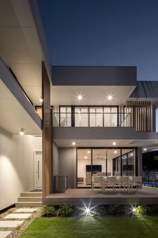 A dusk exterior view of a contemporary multi-level residence centred on a covered outdoor entertaining area. The facade combines crisp white and dark grey volumes, extensive black-framed glazing, vertical timber-look screening, recessed downlights, and a broad timber deck with an outdoor dining setting. A stepped path, gravel garden and lawn occupy the foreground, while an illuminated interior living/dining area is visible through the glass.