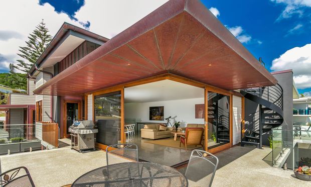Exterior view of a contemporary multi-level home with a large cantilevered copper-toned roof canopy over an open terrace. Floor-to-ceiling timber-framed glazing reveals a bright living room with sofas and armchairs, while a black spiral staircase rises along the right side. The terrace includes an outdoor dining setting, barbecue, glass balustrades, and planted pots. Timber cladding, metal elements, and strong indoor-outdoor connection are visible.