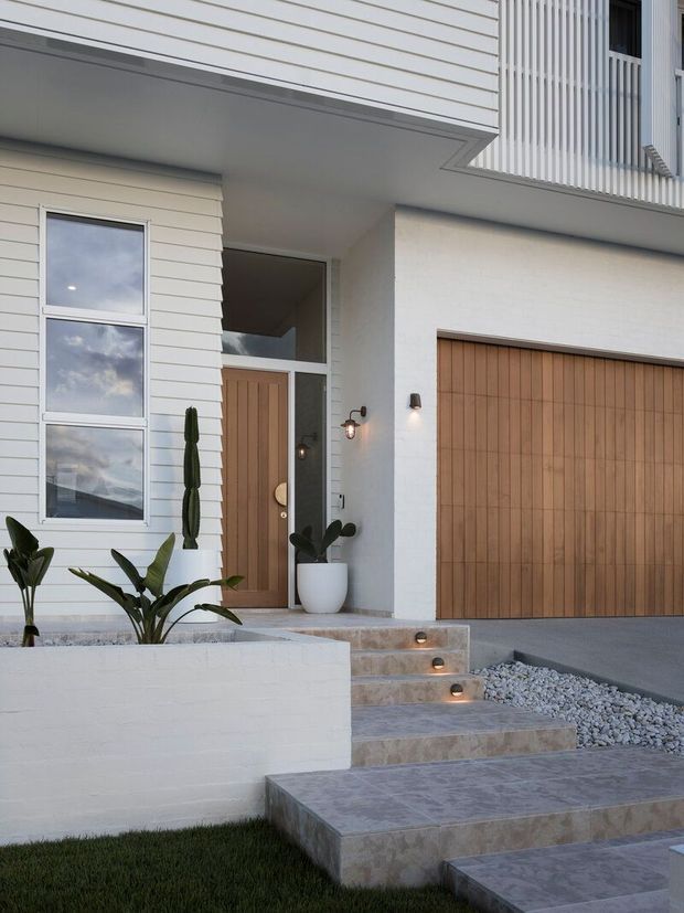 A contemporary residential facade viewed from the front, featuring white horizontal weatherboard cladding, a white textured masonry wall, warm vertical timber garage door and entry door, and a balcony with slim vertical white screening. Broad tiled steps lead to the recessed entrance, bordered by white gravel and a raised white planter with sculptural greenery.