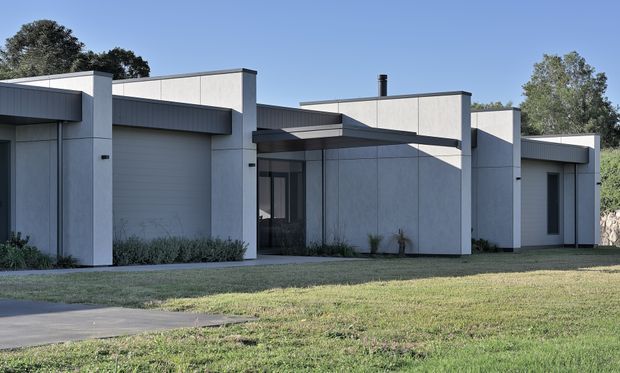 A low-rise contemporary residential facade with a strong horizontal composition, flat roof planes, projecting rectangular forms, and a restrained palette of light grey concrete-like walls with dark charcoal cladding and trim. The frontage includes an integrated garage, recessed glazed entry, narrow vertical windows, and landscaped lawn in the foreground. Mature trees and a clear sky surround the building.