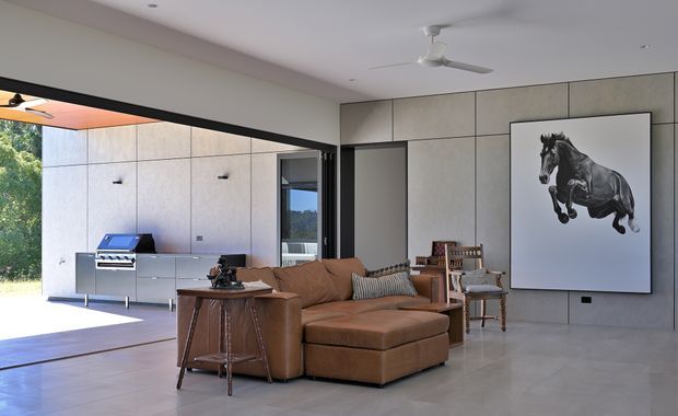 Open-plan contemporary living room with a large brown sectional sofa, timber side table, and occasional chair. The room has expansive sliding or folding glazing opening toward an outdoor kitchen area, large-format pale grey wall panels, polished pale flooring, recessed ceiling lights, and a white ceiling fan. A large monochrome horse artwork forms a feature on the wall.
