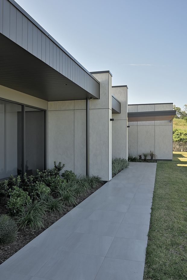 Exterior view of a contemporary single-storey building with sharp geometric volumes, pale grey panelled walls, dark metal soffits and fascia, and large dark-framed glazing. A broad tiled concrete walkway runs alongside low landscaped planting and a lawn, leading toward a covered entrance or secondary volume.