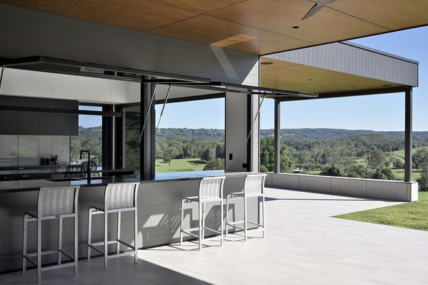 A contemporary open-plan kitchen and entertaining area opening onto a broad covered terrace, with a long island bench and bar stools in the foreground. Large glazed openings frame expansive green rolling hills, while slim dark window frames, exposed structural posts, and a timber-lined ceiling create a refined indoor-outdoor connection. The terrace has pale hard paving and a built-in edge bench or planter.