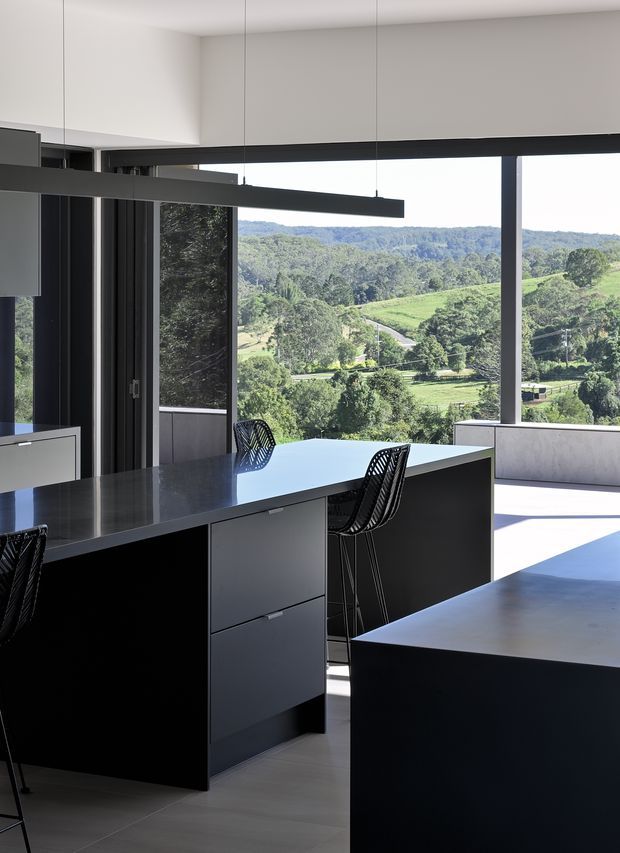 A sleek, contemporary kitchen with a dark island and cabinetry, pale stone or solid-surface benchtops, integrated appliances, and black-framed floor-to-ceiling glazing. Minimal black linear pendant lights hang above the island. The open kitchen overlooks a lush rural, rolling-hills landscape, with strong natural daylight creating a clean, high-contrast interior.