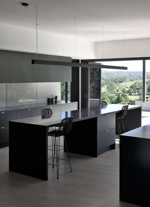 A spacious, minimalist contemporary kitchen with dark charcoal cabinetry, a large central island with integrated seating, and a long linear black pendant light. Pale tiled flooring and white walls provide contrast, while extensive black-framed glazing opens the kitchen to a green rural landscape. The space is clean-lined, refined, and strongly connected to the outdoors.