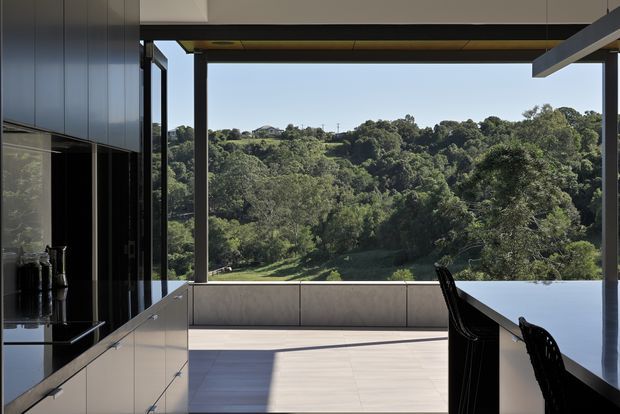Contemporary open-plan kitchen and dining area with dark integrated cabinetry, a glossy island, and a long dining or breakfast counter. Full-height openings frame a lush, wooded hillside, while a deep covered overhang and slim dark structural posts create a strong indoor–outdoor connection. The space is bright, refined, and minimalist with a neutral palette and clean geometric lines.