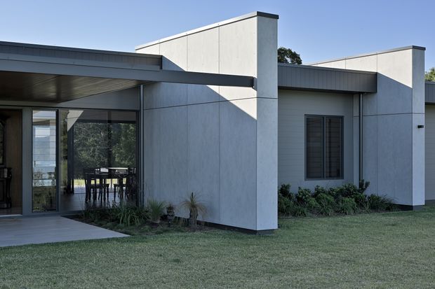 Exterior view of a low-slung contemporary house with crisp geometric volumes, pale smooth concrete or rendered wall panels, dark metal roof edges and framing, and extensive glazed openings beneath a broad covered terrace. The landscaped foreground includes a lawn and low planting, while a dining area is visible through the glazing.