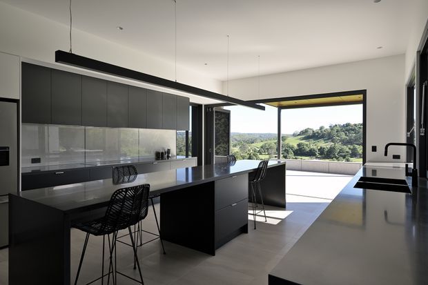 A spacious contemporary kitchen with matte dark cabinetry, a large central island with seating, integrated appliances, and a pale stone or composite benchtop. Full-height glazing and open sliding doors connect the kitchen to a broad terrace, with a green rolling landscape visible beyond. Slim black-framed openings, pendant lighting, and a clean open-plan layout reinforce the modern character.