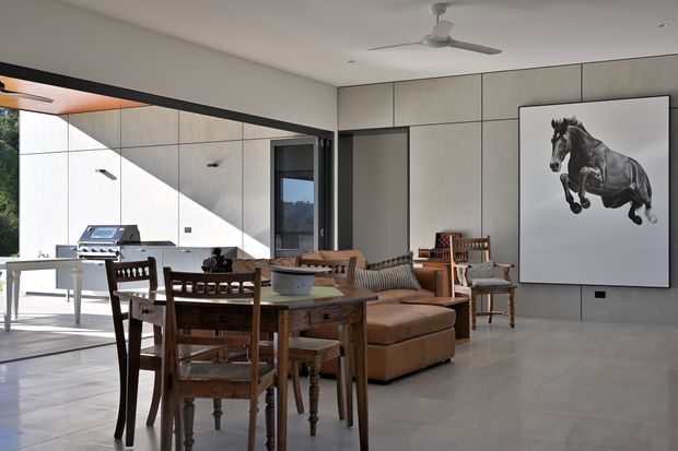 A bright, open-plan contemporary living and dining area with a timber dining table and chairs in the foreground, a large brown upholstered sofa, and additional seating arranged around a low coffee table. Full-height glazed openings connect the interior to an outdoor barbecue area. The room features large pale grey concrete-look wall panels, a smooth light floor, dark-framed glazing and doors, recessed lighting, and a ceiling fan. A large monochrome horse artwork provides a focal point.