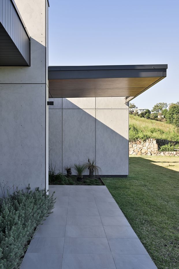A modern residential exterior viewed along a tiled pedestrian path. The facade uses large smooth grey concrete panels with visible joint lines, a dark metal-clad projecting roof edge, and a warm timber-lined soffit beneath a broad cantilever. Low landscaping borders the path, with lawn, stone retaining walls, and vegetation beyond. Strong angular shadows emphasize the minimalist geometric form.