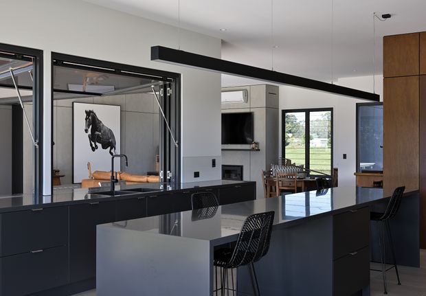 A contemporary open-plan kitchen with a large dark island and integrated seating, viewed toward an adjacent dining and living area. The kitchen features matte black cabinetry, a dark reflective benchtop, black-framed glazed openings, a long linear pendant light, and a tall timber-toned storage wall. A fireplace and television wall are visible beyond the kitchen, with abundant natural light through the glazing.