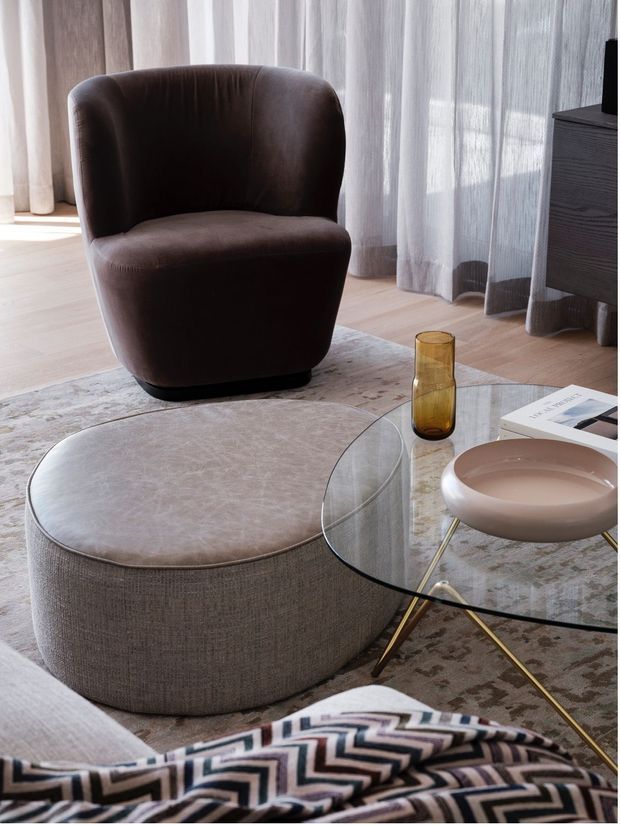 A refined living room vignette featuring a deep brown upholstered lounge chair, a large rounded upholstered ottoman, and a clear glass oval coffee table with slender gold-toned legs. Sheer floor-to-ceiling curtains filter daylight beside dark timber cabinetry, while a muted patterned rug, ceramic bowl, amber glass, and geometric throw add understated decorative detail.