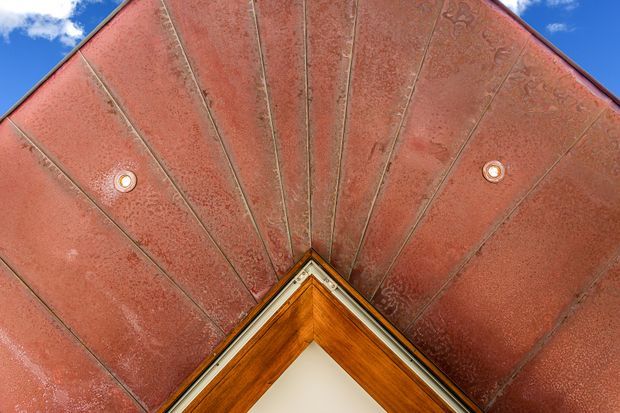 Upward-looking view of a peaked entrance canopy or porch soffit. The red textured ceiling is divided into radiating panels with narrow metal seams and recessed spotlights, converging above a timber-framed triangular doorway or gable detail. Bright blue sky is visible at the edges.