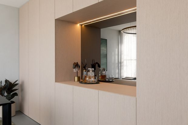 Built-in light timber cabinetry surrounds a recessed display and drinks niche. The niche has warm integrated LED strip lighting, a dark timber or painted interior backing, mirrored rear surface, and styled glassware and bottles. A glimpse of a nearby living space and sheer-curtained window is visible in the reflection. The overall design is clean, contemporary, and warm.