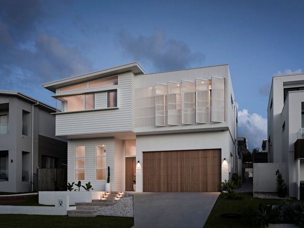 Contemporary two-storey residential house photographed at dusk, featuring a light rendered and horizontal-clad facade, large timber garage door, projecting upper floor, and a prominent vertical screening system across the upper-level windows and balcony. The home sits in a narrow suburban streetscape with a concrete driveway, small landscaped front garden, and neighbouring modern houses.