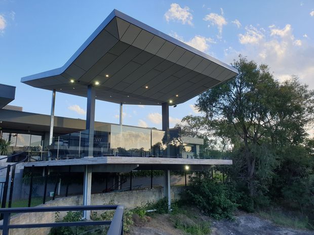 Exterior view of a contemporary multi-level building set among mature vegetation, featuring a dramatic cantilevered flat roof canopy with recessed downlights, slender dark structural columns, extensive glazing, and a terrace or elevated deck. The architecture has strong horizontal lines and an open pavilion-like form.
