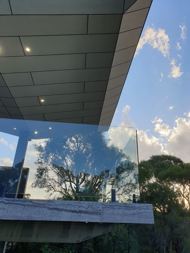 Upward-looking exterior architectural photograph showing a cantilevered, dark panel-clad soffit with recessed downlights, a projecting concrete slab, and frameless glass balustrade or glazing. Trees and blue sky are reflected in the glass, with surrounding vegetation visible beyond. The image emphasizes modern building form, sharp geometric lines, and an outdoor elevated platform or terrace.