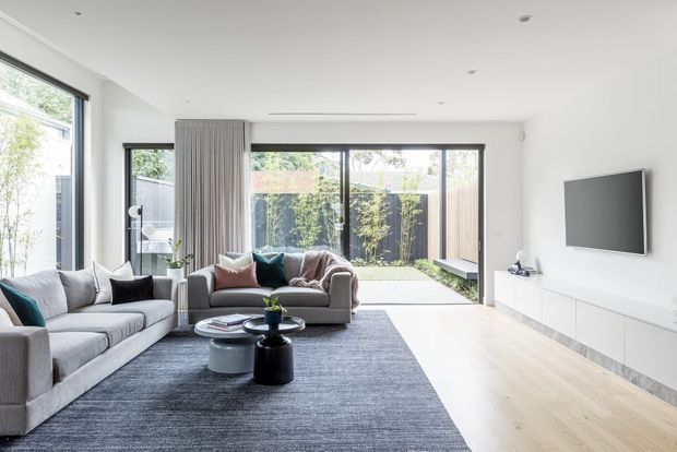 Bright contemporary living room with large grey sofas, a dark textured area rug, nested round coffee tables, and a wall-mounted television above minimalist white built-in cabinetry. Full-height glazing and sliding doors open to a planted courtyard, while pale timber flooring and white walls create an airy, clean interior.