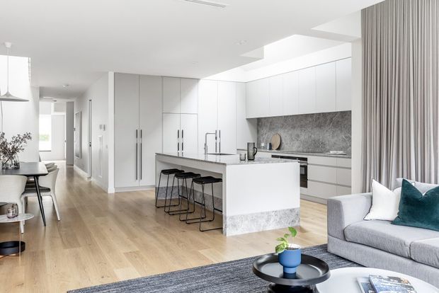 Bright open-plan contemporary kitchen and living area with extensive white flat-panel cabinetry, a large island with seating, grey stone-look benchtops and splashback, pale timber flooring, and a grey sofa beside full-height curtains. A dining area and hallway extend beyond the kitchen.